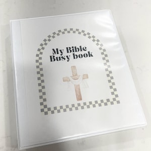 K&ouml;nnte beinhalten: Ein wei&szlig;er Ordner mit einem karierten Rand und dem Text "My Bible Busy Book". Ein Holzkreuz mit einem wei&szlig;en Tuch dar&uuml;ber befindet sich in der Mitte des Ordners.