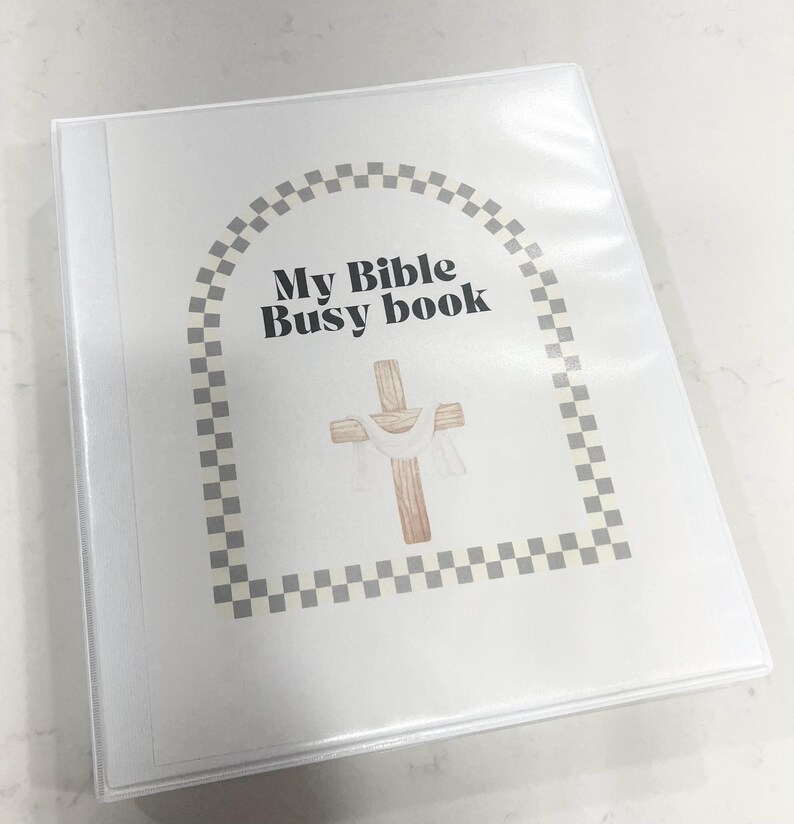 K&ouml;nnte beinhalten: Ein wei&szlig;er Ordner mit einem karierten Rand und dem Text "My Bible Busy Book". Ein Holzkreuz mit einem wei&szlig;en Tuch dar&uuml;ber befindet sich in der Mitte des Ordners.