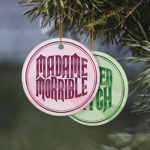 May include: Two round Christmas ornaments hanging from a pine branch. One ornament is pink with the words "MADAME MORRIBLE" in a gothic font. The other is green with the words "EVIL WITCH". Both have gold string hangers.