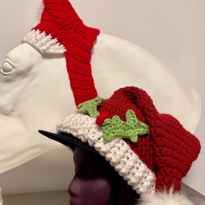 May include: A red and white crocheted Santa hat with a green holly design and a white fur pom-pom. The hat is worn on a mannequin head.