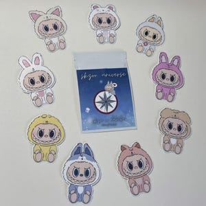 May include: A collection of eight die-cut stickers featuring cartoon characters in various animal-themed onesies, surrounding a central rectangular clear plastic pouch. The pouch displays a blue graphic with the text skzoo universe.