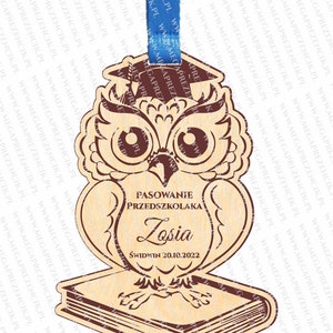 May include: Wooden owl figurine with a graduation cap, holding a book. The owl is brown with red eyes and a brown book. The text on the owl reads "Pasowanie Przedszkolaka Losia Świdwin 20.10.2022".