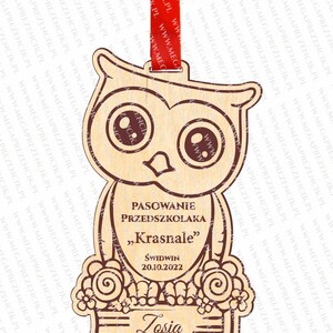 May include: A wooden owl-shaped plaque with a brown outline. The plaque has the text "PASOWANIE PRZEDSZKOLAKA "Krasnale" ŚWIDWIN 20.10.2022" and the name "Zosia" written on it. The plaque is decorated with flowers and a book.