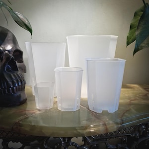 May include: Five clear, geometric plastic plant pots of different sizes. The pots are arranged on a decorative table. A dark skull and greenery are in the background. The pots have a frosted appearance, with a hexagonal design.