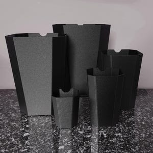 May include: Five black, geometric containers with a textured finish. The containers are of varying sizes, each with a small notch at the top. They are arranged on a speckled surface. The containers are made of a dark material.