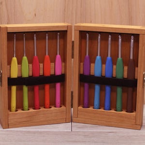 May include: A wooden box with two compartments, each containing a set of colorful crochet hooks. The hooks are arranged in rows and are held in place by a black elastic band.