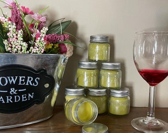 May include: A stack of seven small glass jars with silver lids filled with a green substance. The jars are arranged in a pyramid shape. There is a glass of red wine and a metal bucket with flowers in the background.