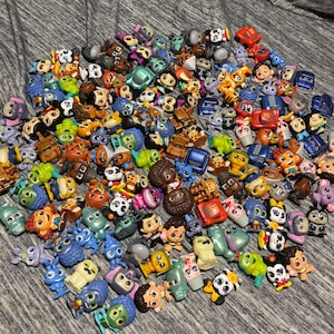Può includere: Una vasta collezione di piccole statuette in plastica colorata di varie forme, tra cui animali, veicoli e figure umane con espressioni diverse, disposte in un mucchio denso e sparso su una superficie in tessuto grigio.