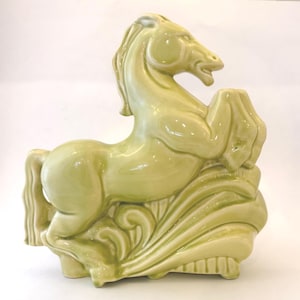 Puede incluir: Figura de caballo de cerámica verde claro en pose encabritada. El caballo está esculpido con detalles y descansa sobre una base decorativa con un diseño de olas. La figura mide aproximadamente 25 cm de altura.