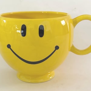 May include: A yellow ceramic mug with a smiley face design. The mug features a large, round body, a curved handle, and a small base. The smiley face has black eyes and a curved black mouth.