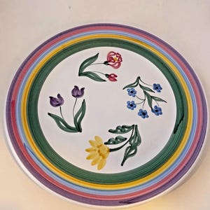 May include: A round white ceramic plate with a rainbow striped border. The plate features hand-painted floral designs, including tulips, forget-me-nots, and a sunflower. The border has stripes of purple, pink, yellow, and green.