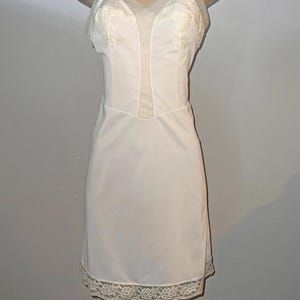 Peut inclure: Une robe slip blanche avec des bretelles réglables et une délicate bordure en dentelle à l'ourlet. Le corsage présente des panneaux transparents et des détails en dentelle. La robe est présentée sur un mannequin.