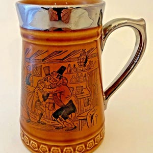 May include: Brown ceramic beer stein with a silver-colored rim and handle. The stein features a detailed illustration of two men embracing in a pub setting. The base has a decorative pattern.