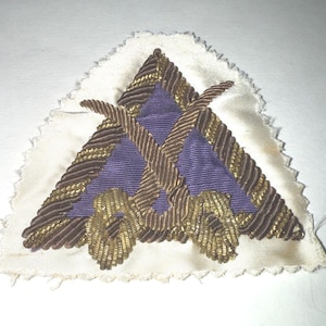 Peut inclure: Un écusson triangulaire avec une base en tissu blanc et un motif décoratif. Le motif présente un centre violet avec des détails brodés dorés et bruns, comprenant des éléments croisés et des ornements. Les bords de l'écusson sont inachevés.