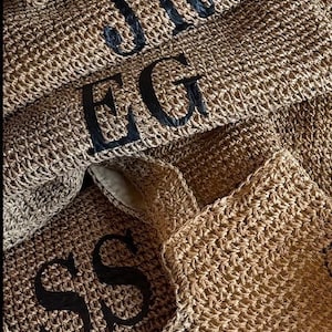 May include: A close-up of a stack of woven straw bags with black embroidered initials. The initials are "SS", "EG", "JR", "RS", and "BW".