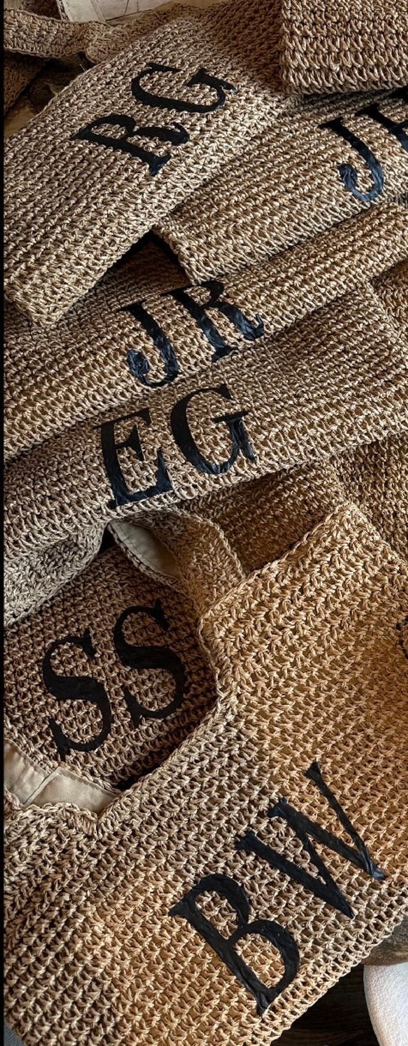 May include: A close-up of a stack of woven straw bags with black embroidered initials. The initials are "SS", "EG", "JR", "RS", and "BW".
