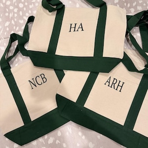 May include: Three canvas tote bags with cream-colored bodies and dark green accents. Each bag has a dark green trim and handles. The bags are personalized with the initials "HA", "NCB", and "ARH" in black block letters.
