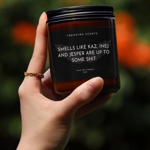 May include: A brown glass candle with a black label that reads "Trending Scents" and "Smells like Kaz, Inej and Jesper are up to some shit." The candle is 100% soy and is 8 oz.