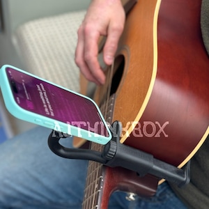 May include: A black phone holder attached to a brown acoustic guitar. The phone is light blue and has lyrics displayed on the screen.