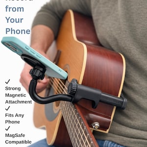 May include: A black phone mount attached to an acoustic guitar, holding a light blue phone. Text on the image reads "Play, Learn & Record from Your Phone." Features include a strong magnetic attachment, fits any phone, and is MagSafe compatible.