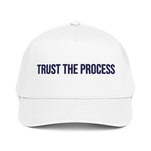 Peut inclure: Casquette de baseball blanche avec une visière incurvée. L'inscription "TRUST THE PROCESS" en bleu marine est imprimée sur le devant. La casquette a une coupe structurée et un bouton sur le dessus.