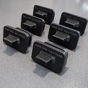 May include: Six black rectangular automotive parts with a raised handle and the text "BMW MINI SOLUTIONS" in white. The parts are arranged on a gray surface, with the text clearly visible on each.