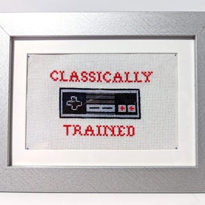 May include: A framed cross-stitch artwork with a silver frame. The artwork features a black and white controller design with the words "CLASSICALLY TRAINED" in red. The background is white.