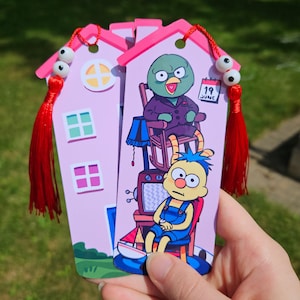 May include: Two pink house-shaped bookmarks with cartoon characters. Each bookmark has a pink roof, windows, and a tassel. One bookmark features a green bird character in a purple suit, and the other features a yellow character in blue overalls.