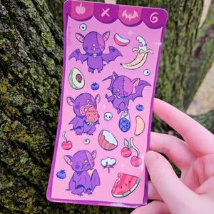 May include: A pink sticker sheet featuring purple cartoon bats with various fruits and a skull. The bats have large wings and are depicted with different expressions. Other illustrations include a banana, avocado, and watermelon. The sticker sheet is held by a person.