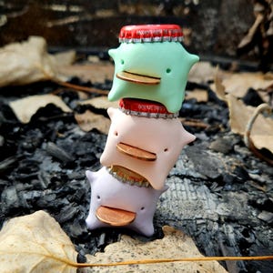 May include: Three pastel-coloured sculptures stacked on top of each other, each resembling a small, smiling creature with a coin as a beak. A red bottle cap sits between each sculpture. The background is a dark, textured surface, possibly charcoal.