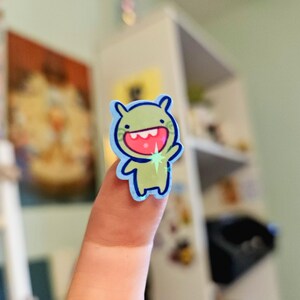 May include: A green monster sticker with a big smile and a blue outline. The sticker has a sparkly star on its hand.