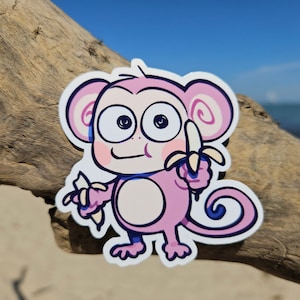 May include: A pink cartoon monkey with big eyes and a smile, holding a banana in each hand. The monkey is standing on a sandy surface.