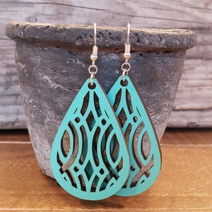 May include: Teal teardrop earrings with a decorative cut-out design. The earrings have silver-colored hooks and are displayed against a rustic wooden surface. The earrings are a stylish accessory.
