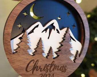 May include: A wooden Christmas ornament with a laser-cut design of a mountain range, trees, a crescent moon, and stars against a black background. The ornament is inscribed with "Christmas 2024".