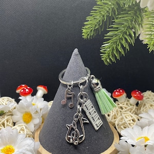 Peut inclure: Porte-clés argenté sur le thème de la musique avec un pompon vert. Les breloques comprennent une clé de sol, une croche et des touches de piano. Le porte-clés est présenté sur un support conique gris, entouré de fleurs blanches et de verdure.