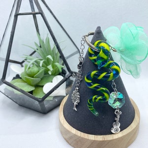 Op de afbeelding: Een blauwe en gele glazen slangbedel met een zilveren ketting en bedels, waaronder een boom, een kristal en een muzieknoten. De bedel wordt getoond op een grijze en houten displaystandaard.