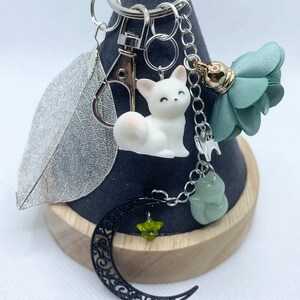 Peut inclure: Un porte-clés argenté avec divers breloques. Les breloques comprennent une figurine de chat blanc, une feuille argentée, un croissant de lune filigrané noir avec une étoile verte, une pierre verte et une fleur en tissu vert.