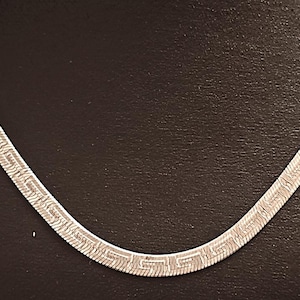 May include: Silver-toned necklace with a flat, herringbone chain design. The necklace features a repeating geometric pattern along its length, creating a textured appearance. The jewelry is displayed against a dark background.