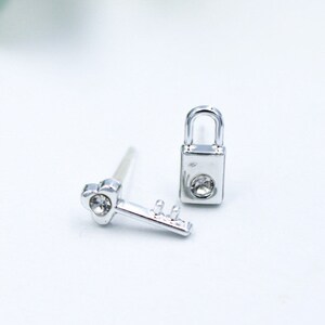 Puede incluir: Un par de pendientes de plata, uno con forma de candado con un strass transparente y el otro con forma de llave con un strass transparente.