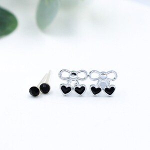 May include: A set of two pairs of earrings. One pair features a silver bow with two black heart charms. The other pair features a single black gemstone.