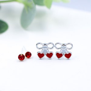 May include: A set of three pairs of earrings. Two pairs feature a silver bow with two red hearts. The third pair features a single red rhinestone.