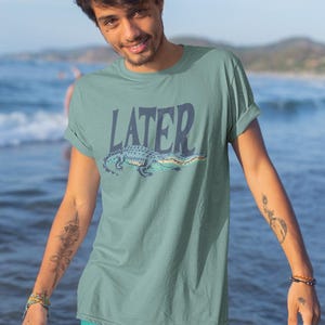 K&ouml;nnte beinhalten: Ein tealfarbenes T-Shirt mit dem Wort "LATER" &uuml;ber einem Cartoon-Alligator. Der Alligator ist blau und gr&uuml;n. Die Person auf dem Bild tr&auml;gt eine tealfarbene Shorts und steht vor einem Gew&auml;sser.