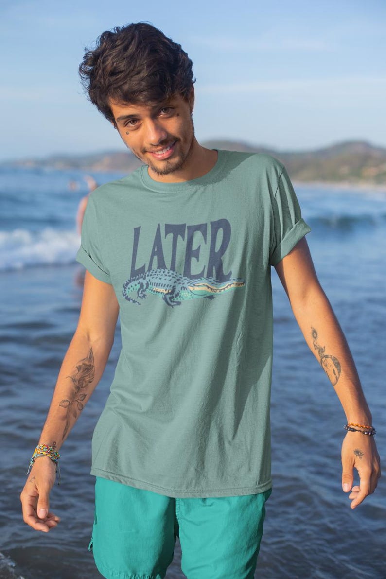 K&ouml;nnte beinhalten: Ein tealfarbenes T-Shirt mit dem Wort "LATER" &uuml;ber einem Cartoon-Alligator. Der Alligator ist blau und gr&uuml;n. Die Person auf dem Bild tr&auml;gt eine tealfarbene Shorts und steht vor einem Gew&auml;sser.