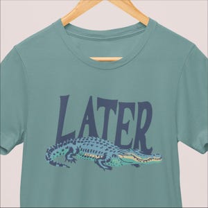 K&ouml;nnte beinhalten: Ein teal-farbenes T-Shirt mit einer Illustration eines blauen Alligators und dem Wort "LATER" in Marineblau. Der Alligator hat ein blaues und gr&uuml;nes Muster mit gelben Akzenten. Das T-Shirt h&auml;ngt an einem Holzkleiderb&uuml;gel.