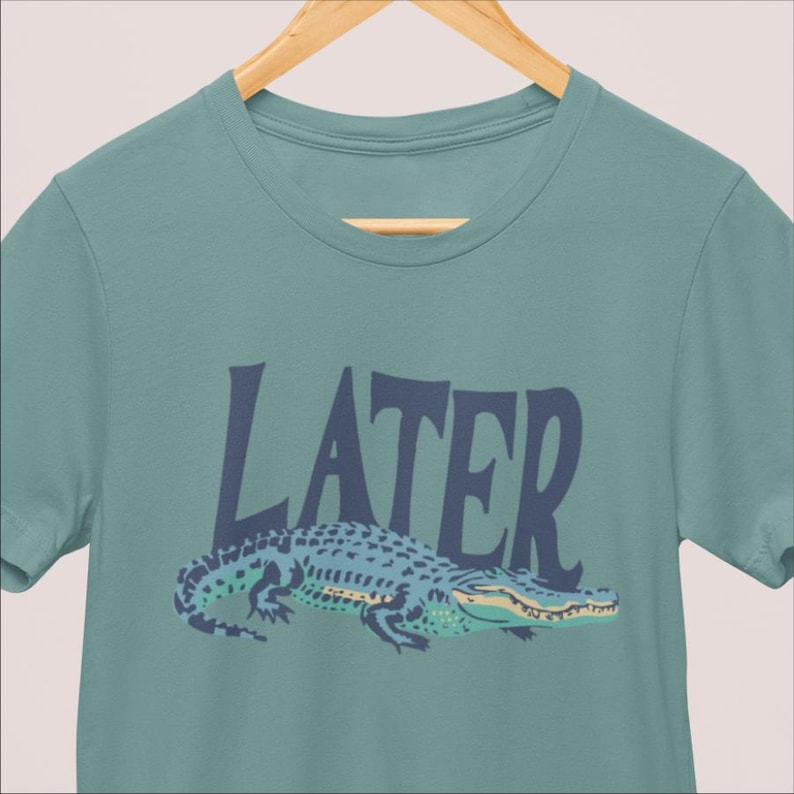 K&ouml;nnte beinhalten: Ein teal-farbenes T-Shirt mit einer Illustration eines blauen Alligators und dem Wort "LATER" in Marineblau. Der Alligator hat ein blaues und gr&uuml;nes Muster mit gelben Akzenten. Das T-Shirt h&auml;ngt an einem Holzkleiderb&uuml;gel.