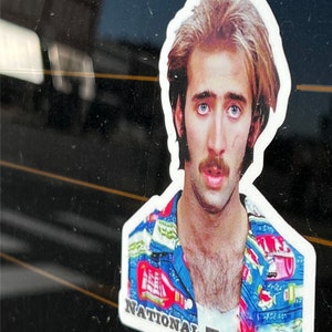 May include: A sticker featuring a close-up portrait of Nicolas Cage wearing a blue and red patterned shirt. The sticker has the text "NATIONAL TREASURE" at the bottom.
