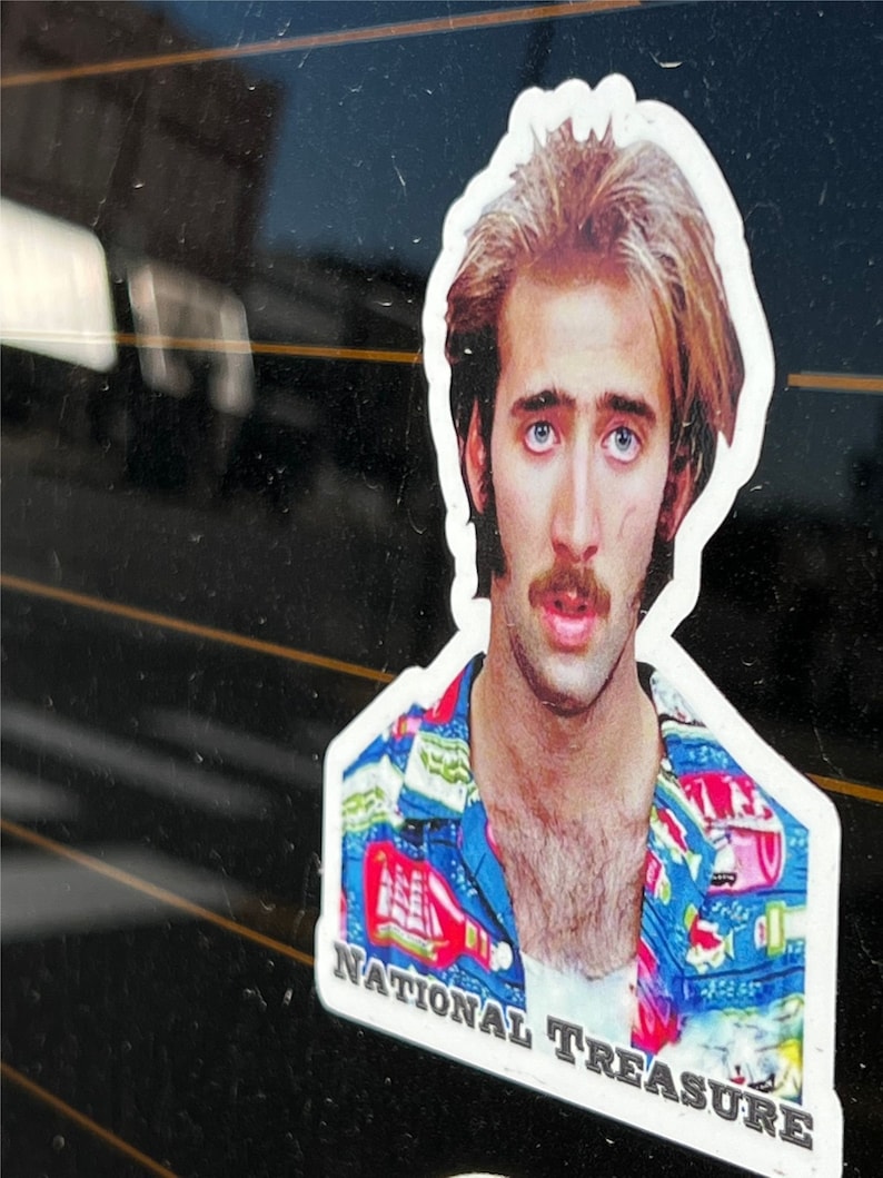 May include: A sticker featuring a close-up portrait of Nicolas Cage wearing a blue and red patterned shirt. The sticker has the text "NATIONAL TREASURE" at the bottom.