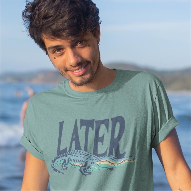 K&ouml;nnte beinhalten: Ein tealfarbenes T-Shirt mit dem Wort "LATER" in marineblauen Blockbuchstaben und einer Cartoon-Alligator-Illustration in Teal, Marine und Gelb. Das Shirt wird von einer Person getragen.