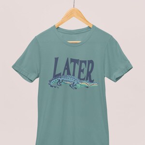 K&ouml;nnte beinhalten: T&uuml;rkisfarbenes T-Shirt mit dem Wort &bdquo;LATER&ldquo; in dunkelblauen Gro&szlig;buchstaben &uuml;ber einer blau-gr&uuml;nen Alligatoren-Illustration. Das Shirt h&auml;ngt an einem Holzb&uuml;gel.