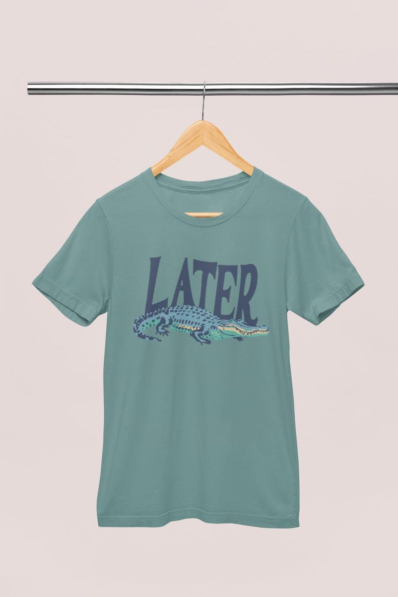 K&ouml;nnte beinhalten: T&uuml;rkisfarbenes T-Shirt mit dem Wort &bdquo;LATER&ldquo; in dunkelblauen Gro&szlig;buchstaben &uuml;ber einer blau-gr&uuml;nen Alligatoren-Illustration. Das Shirt h&auml;ngt an einem Holzb&uuml;gel.